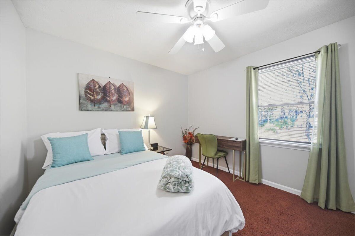 A hotel room with a bed, a desk, a chair, a ceiling fan, a window with curtains, and a painting on the wall.