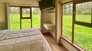 A bedroom with a bed, a TV mounted on the wall, and large windows offering a view of a grassy area.