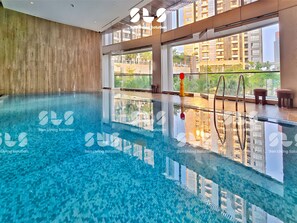 A modern indoor swimming pool with clear water, large windows, and a view of surrounding buildings. - Agio By SanLiving - 2BR - HubLife Mall (Jakarta)