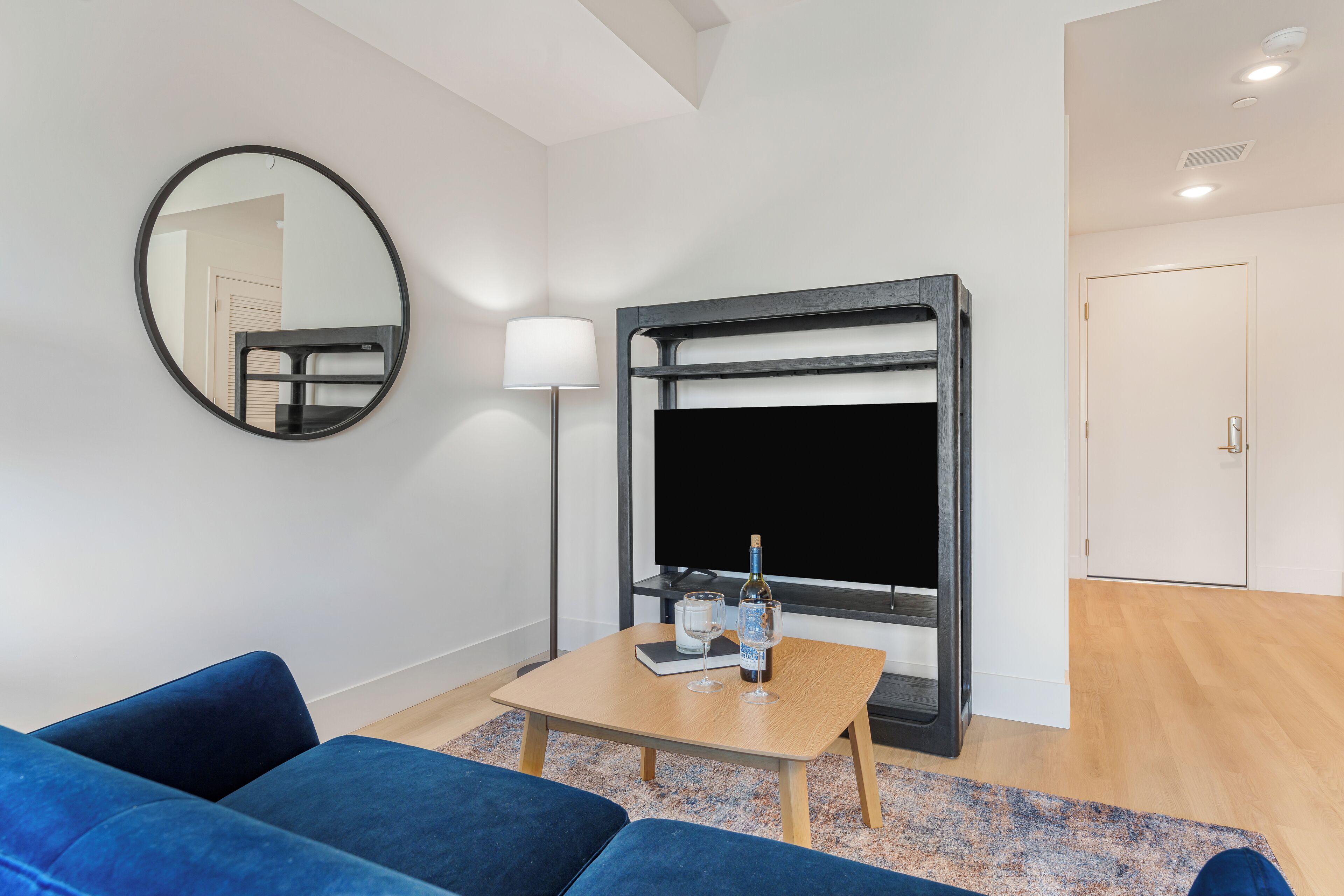 A modern room with a sofa, a small table with a bottle and glass, a TV, a mirror, and a door.