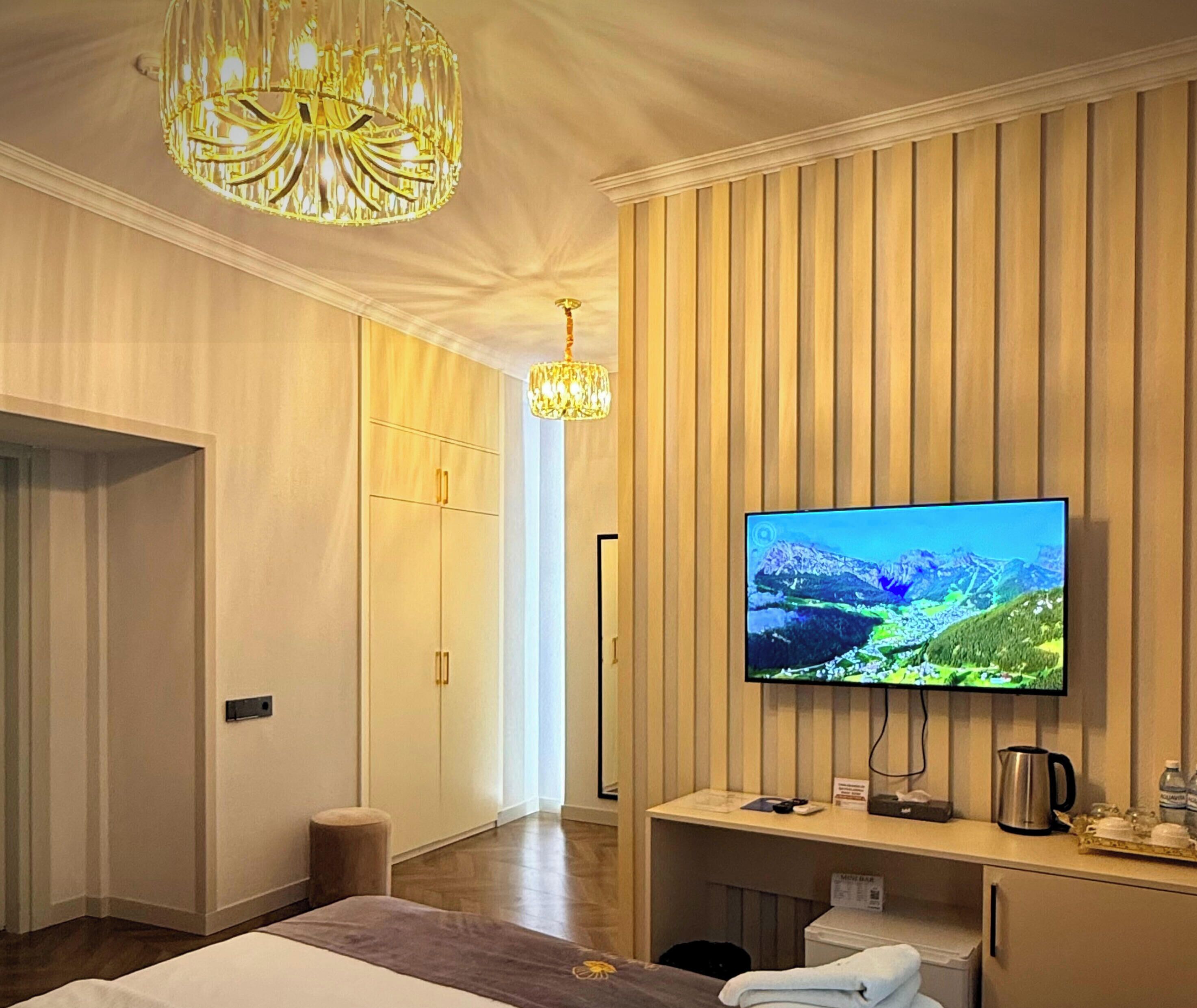 A hotel room with a large flat-screen TV mounted on a wooden wall, a desk with a coffee maker, and a chandelier.