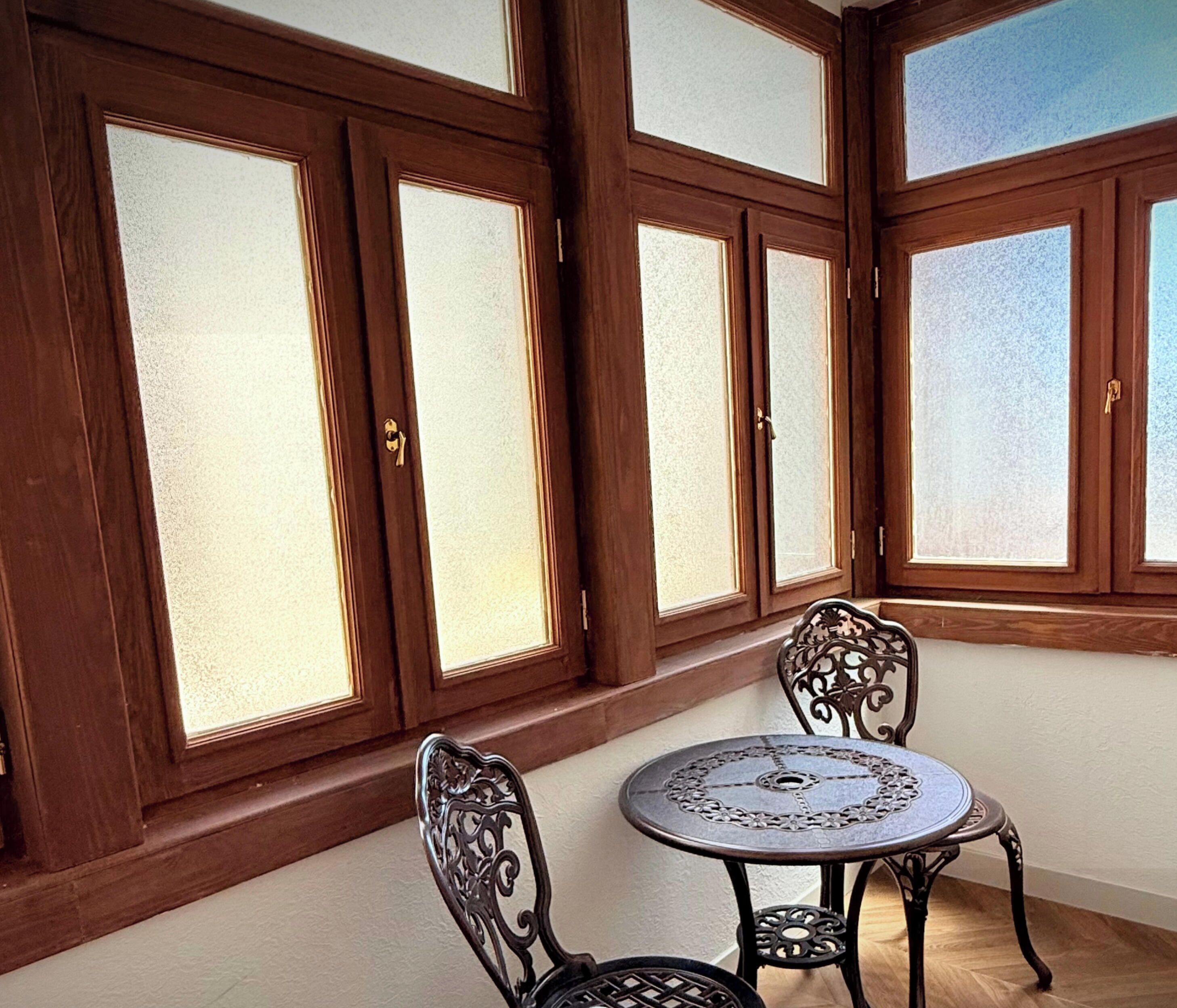 A room with a round table and two chairs, large wooden-framed windows, and a white wall.