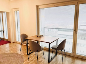 Una habitación con una mesa de madera y cuatro sillas, ventanas amplias con persianas y vistas a un paisaje urbano.
