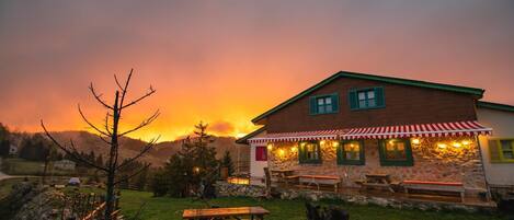 A rustic stone building with a red and white striped awning, outdoor seating, and a picnic table in a grassy area with a sunset in the background.
