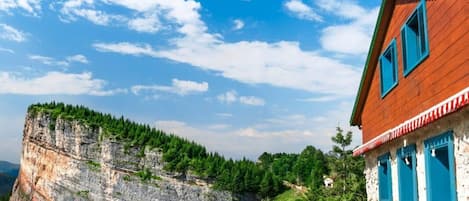 Una casa rustica con facciata in legno e persiane blu, con vista su una scogliera e una vegetazione rigogliosa.