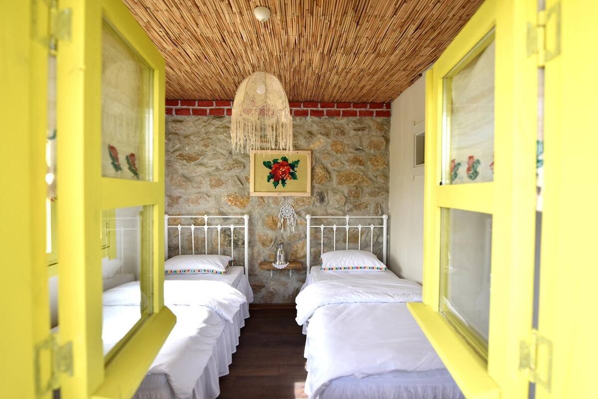 Une chambre avec deux lits, un mur en pierre, un plafond en chaume et un tableau représentant des fleurs.