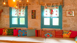 A cozy room with a wooden ceiling, two windows with blue shutters, and a colorful sofa with various patterned cushions. - Kayaüstü Dağ Evi (Duzkoy)