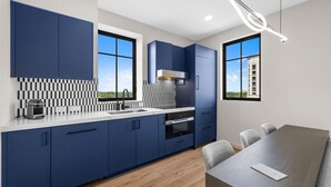 Uma cozinha moderna com armários azuis, bancada branca e forno embutido. Há uma mesa de jantar com cadeiras e vista para um edifício pela janela.