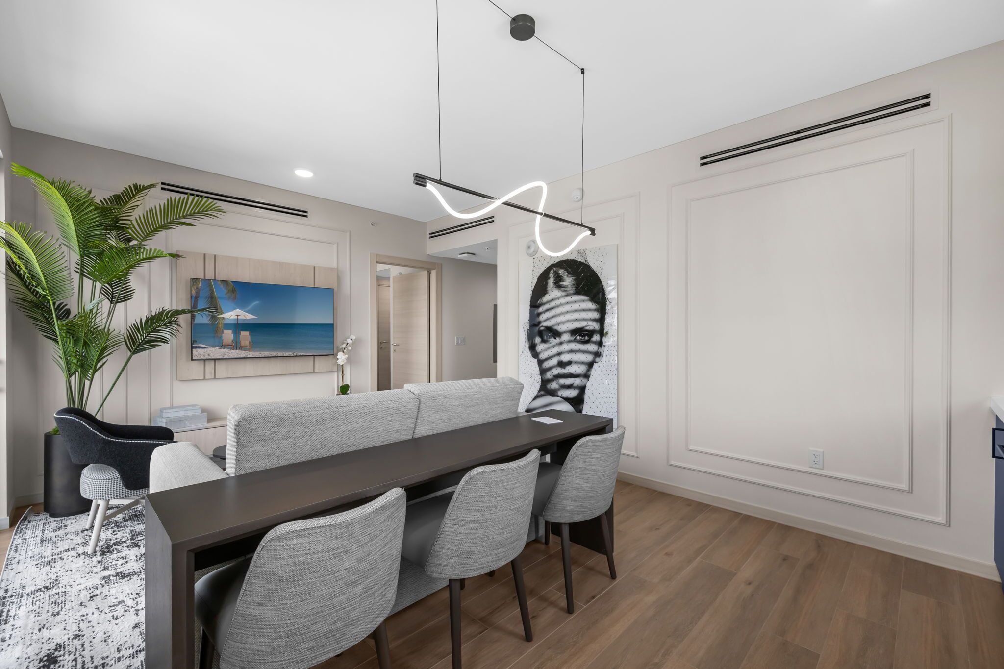 Un espace de salle à manger moderne avec une longue table, des chaises à revêtement gris, une plante en pot et un tableau représentant une scène de plage.