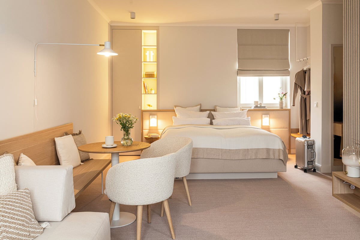 A hotel room with a bed, a sofa, a chair, a bedside table with a lamp, a small table with a vase, and a window with a view.