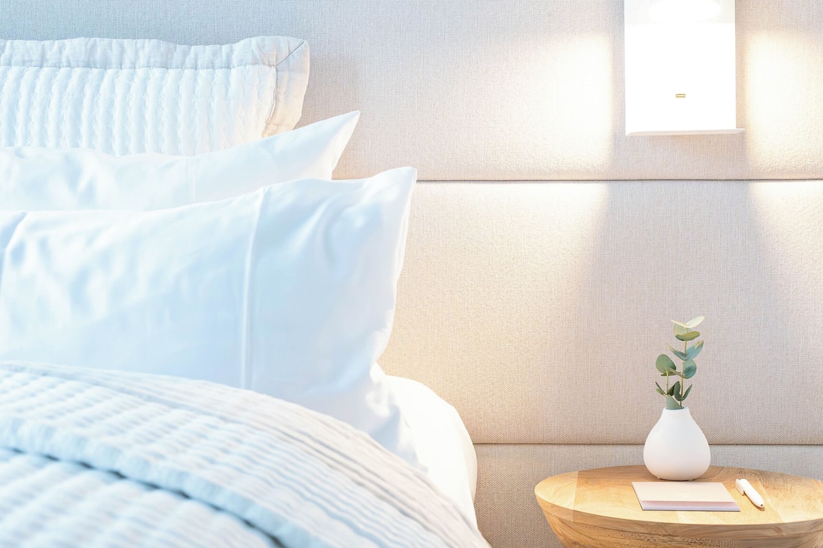 A neatly made bed with white bedding, a round wooden side table with a vase of green leaves, and a wall-mounted light fixture.