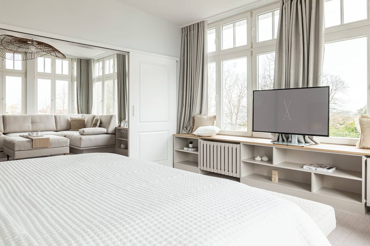 A modern bedroom with a large bed, a TV on a wooden desk, and a view of trees through the windows.