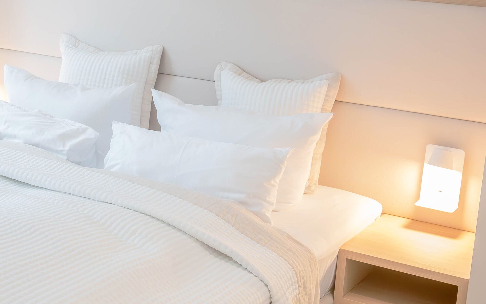 A neatly made bed with white bedding and pillows, a bedside table with a lamp, and a wooden headboard.