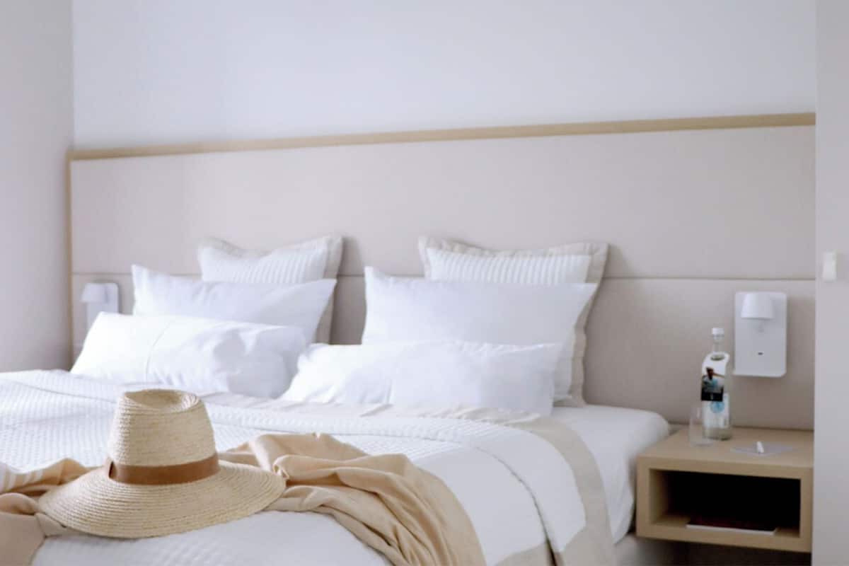 A neatly made bed with white linens and a straw hat on the bed. There is a bedside table with a bottle and a lamp.