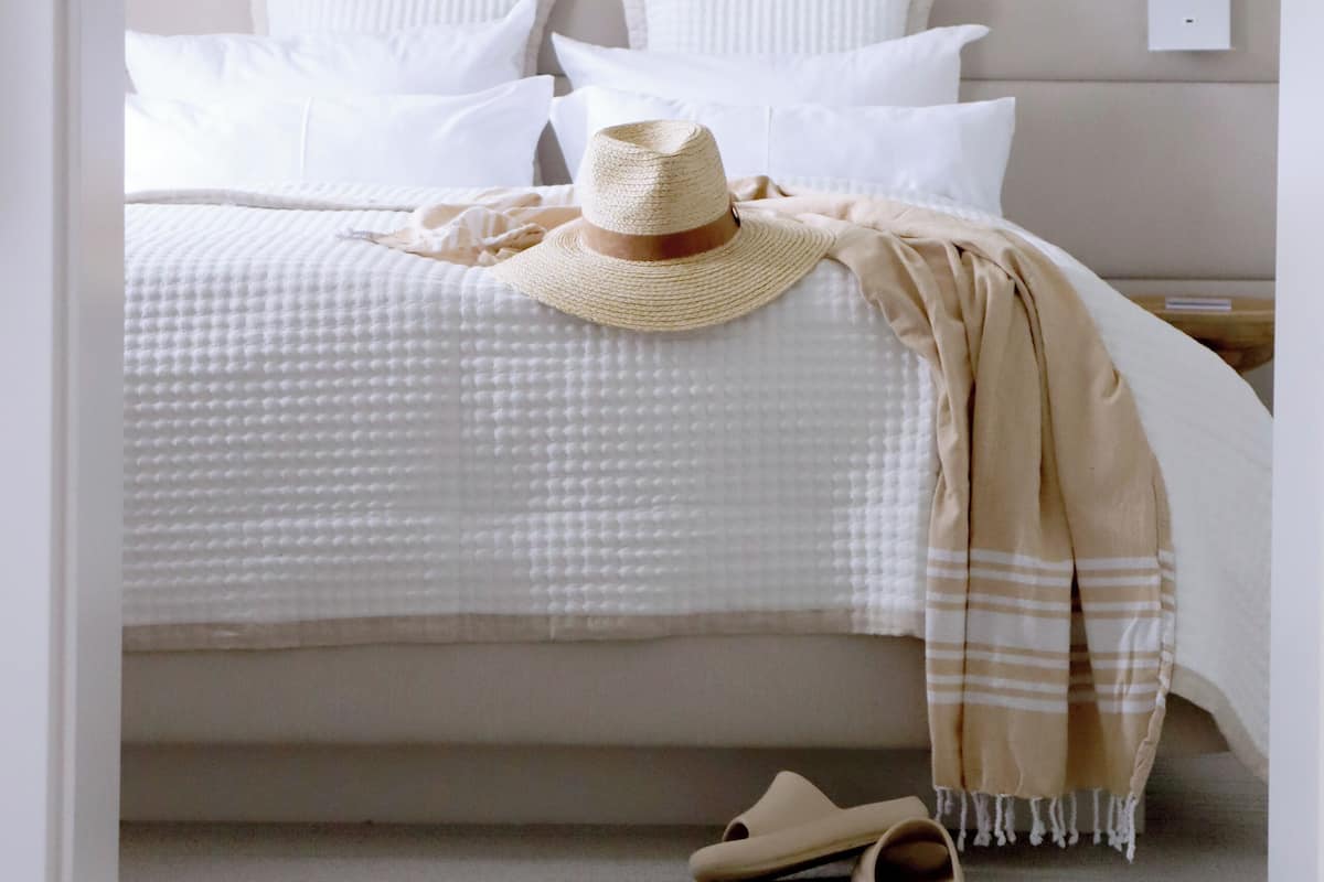 A neatly made bed with white bedding and a beige throw blanket, a straw hat, and a pair of slippers on the floor.