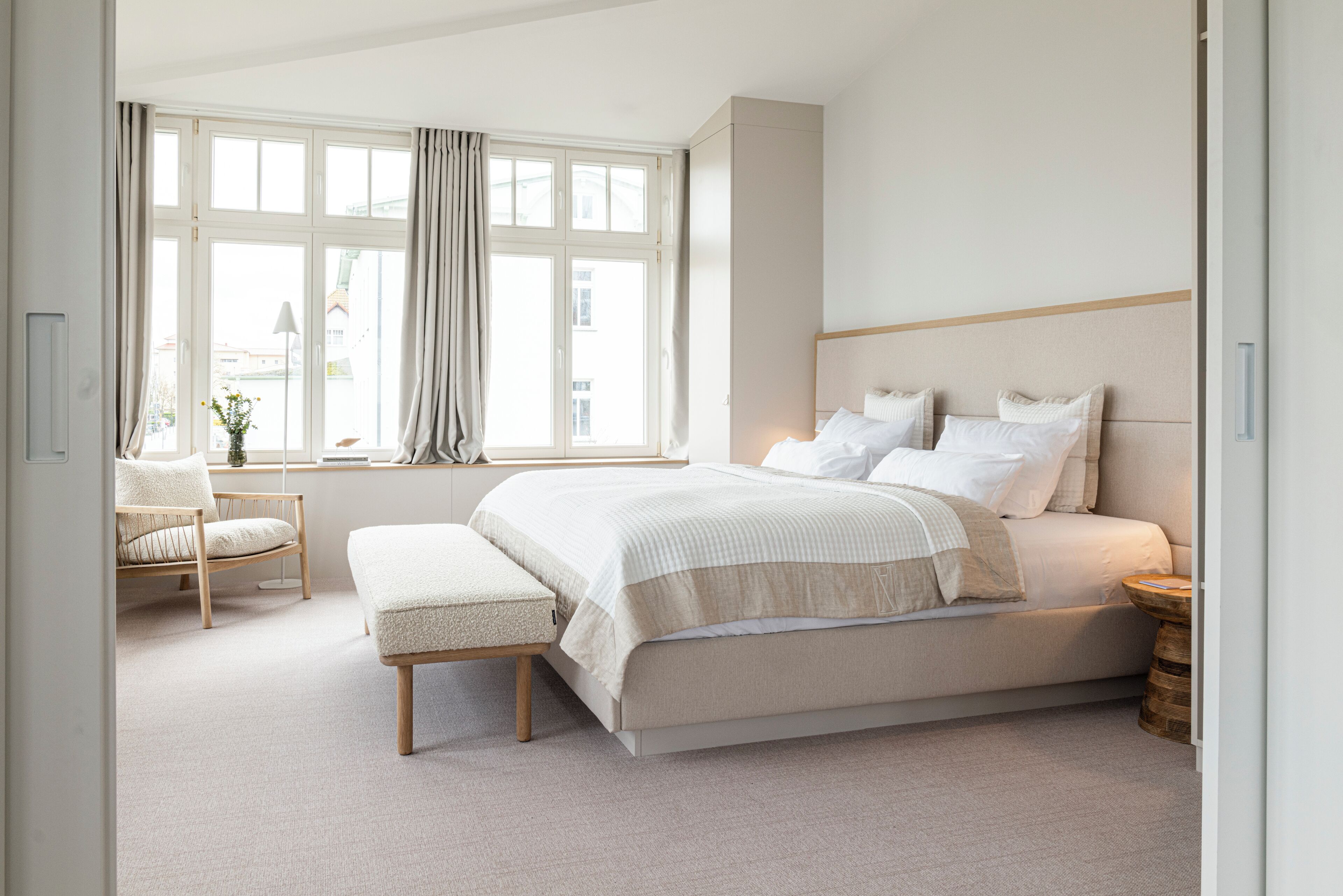A bedroom with a large bed, a window with curtains, a chair, and a bedside table.