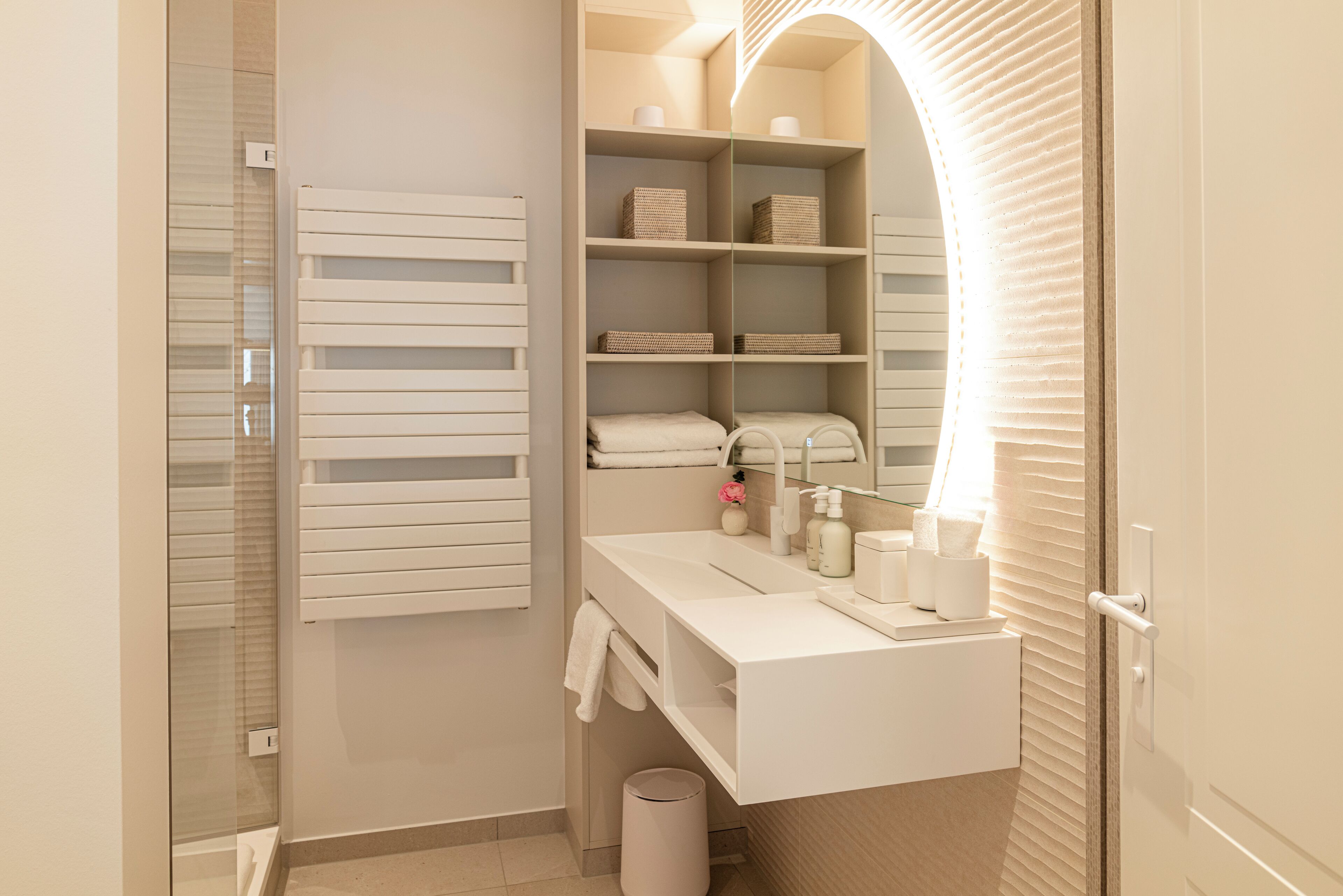 Un baño moderno con cabina de ducha de vidrio, tocador blanco con espejo y estanterías empotradas con cestas de almacenamiento.