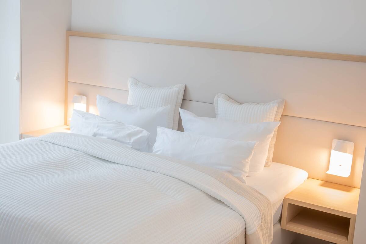 A neatly made bed with white bedding and pillows, a wooden headboard, and bedside lamps.