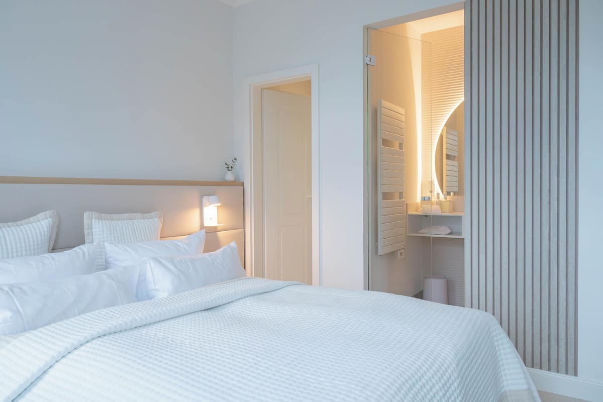 A modern hotel room with a neatly made bed, a bedside lamp, and a view of the bathroom through a glass door.