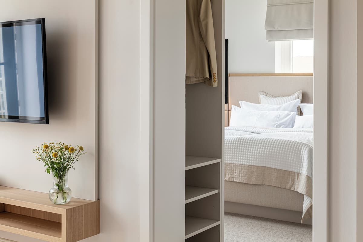 A hotel room with a TV mounted on the wall, a bedside table with a vase of flowers, a bed with white bedding, and a closet with hanging clothes.