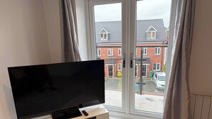 Ein modernes Wohnzimmer mit Flachbildfernseher auf einer weißen Unterhaltungseinheit, großen Fenstern mit Vorhängen und Blick auf eine Wohnstraße.