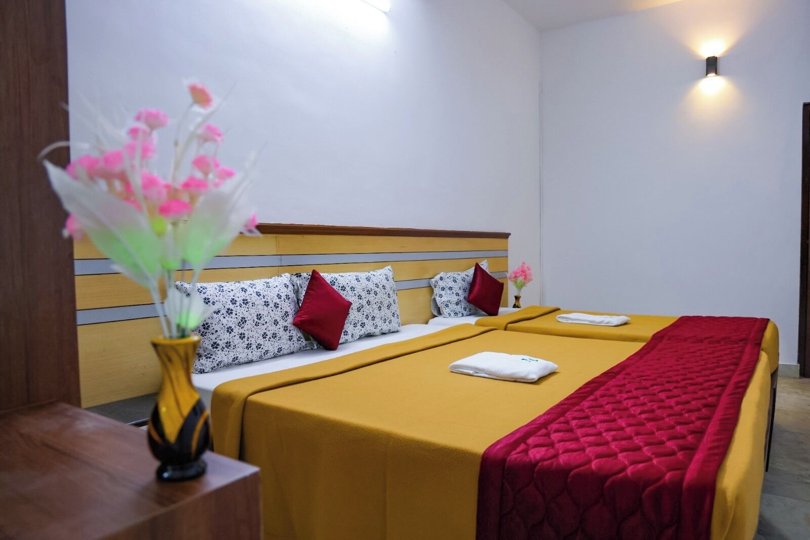 A hotel room with a bed, a bedside table, a vase with flowers, and a wall-mounted light.