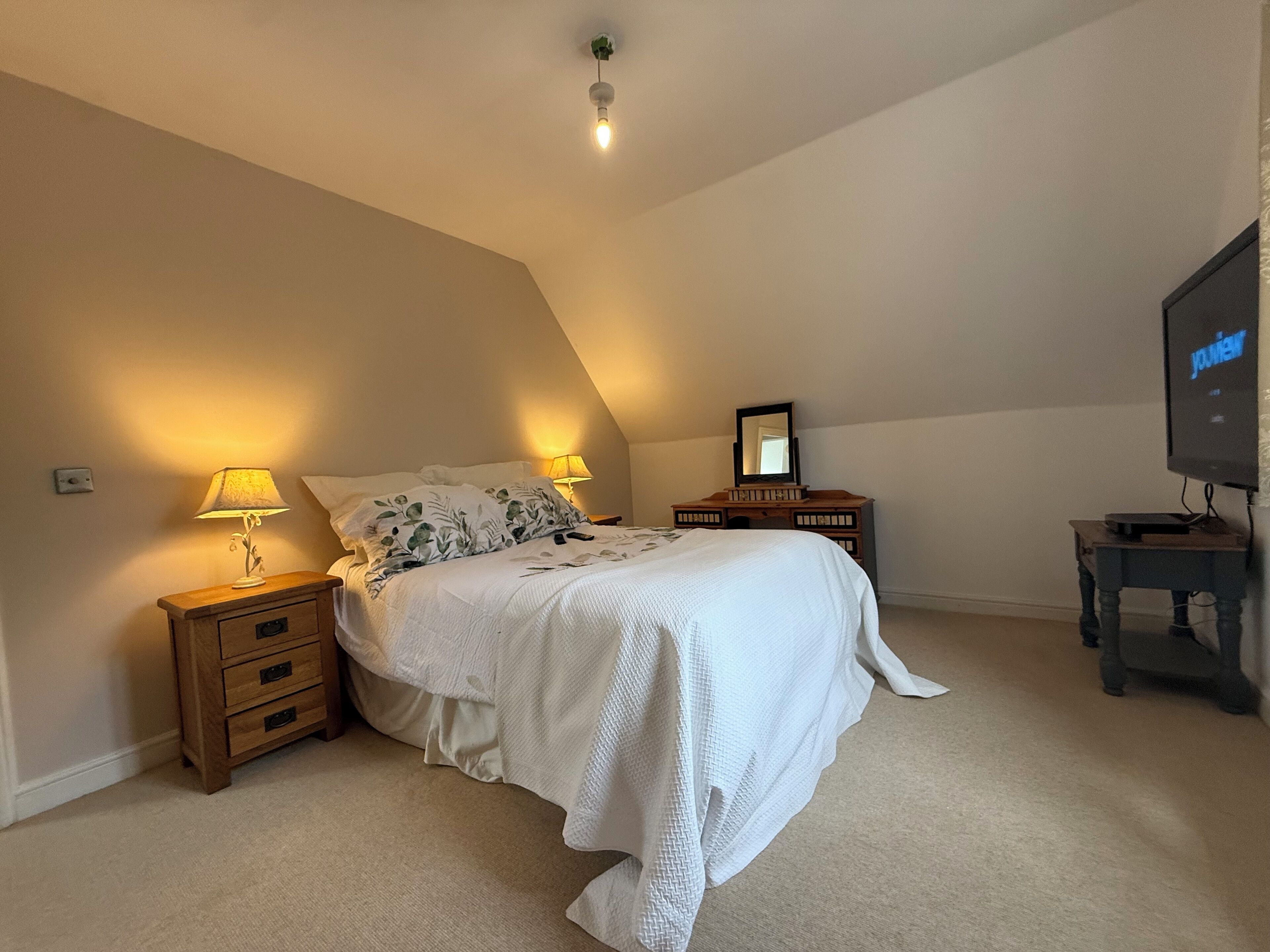 Un dormitorio con cama, dos mesitas de noche, cómoda, espejo y televisor.