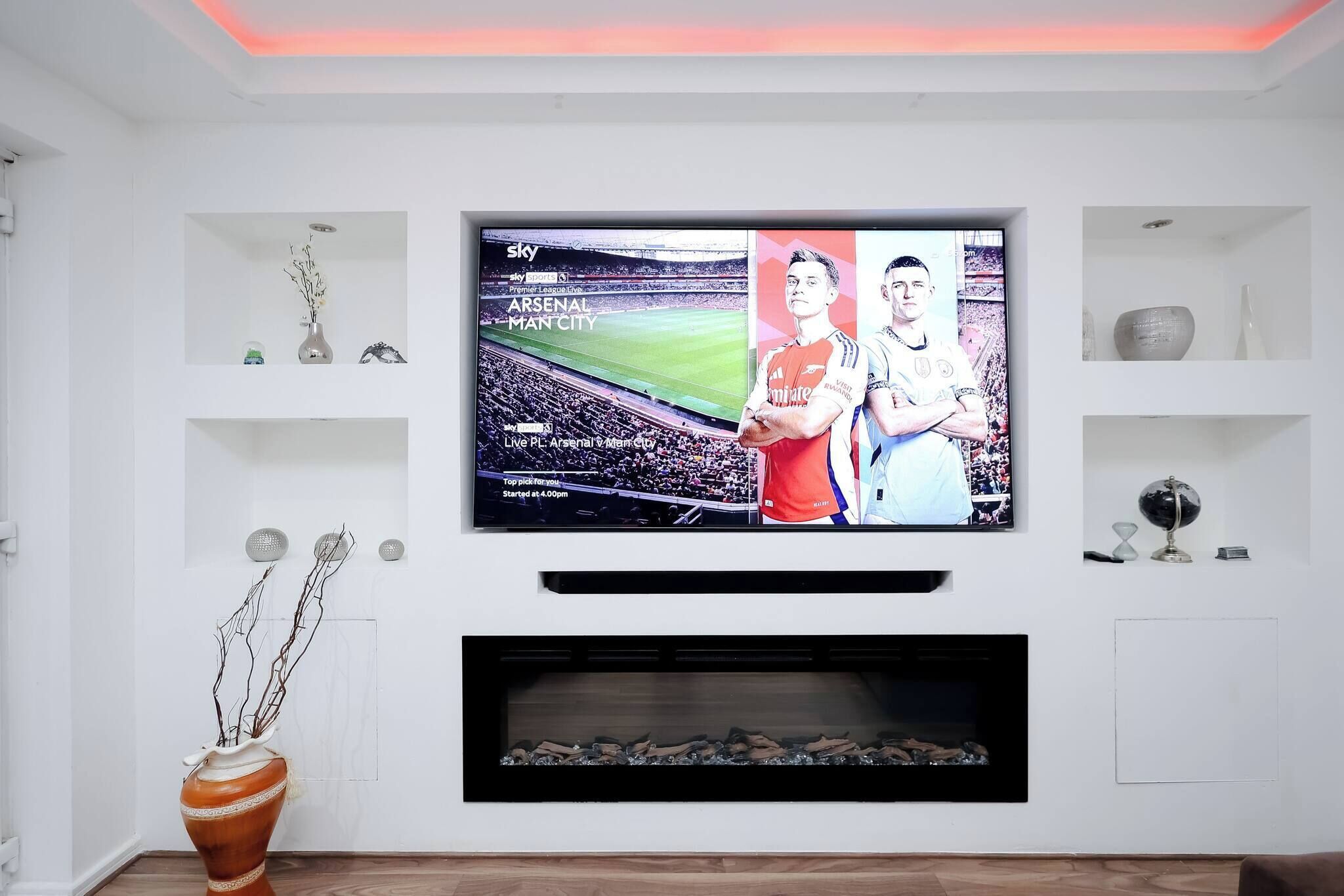 A modern living room with a flat-screen TV mounted on the wall, a fireplace below, and decorative items on shelves.