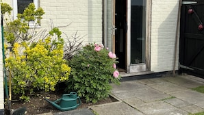 Een wit bakstenen gebouw met een houten deur, een kleine tuin met geel en roze beplanting, en een groene gieter op de grond.