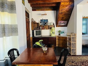 A dining area with a wooden table, chairs, and a plant centerpiece. A television is mounted on the wall, and there are shelves with bottles and a small cabinet. - Sasru Holiday Home (Nuwara Eliya)
