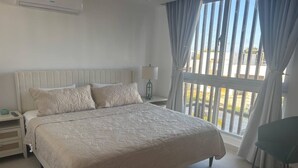 Un dormitorio con una cama grande, una mesita de noche, una lámpara y vistas al exterior a través de una ventana con cortinas.