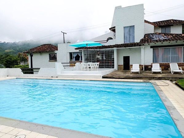 A swimming pool with a covered area and lounge chairs, a white building with a tiled roof, and a mountainous backdrop.