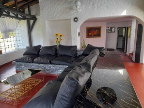 A modern living room with a black leather sofa, a glass coffee table, and a vase with yellow flowers. There is a painting of fruit on the wall and a staircase leading to another room.