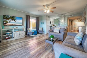 A spacious living room with a large sofa, a coffee table, and a television. There is a kitchen in the background, and a balcony with a view of the ocean. - Port A Condo + Easy Beach Access + Pool + Tennis, Pickeball and Basketball Court (Port Aransas)
