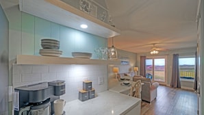 Een moderne keuken met een koffiezetapparaat, theezakjes en koffie, en uitzicht op de woonkamer met een bank en een balkon.