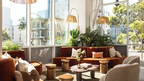 A modern living room with a sofa, coffee table, and wooden stools. Large windows offer a view of the city. - Rooftop Retreat + Spa + Pool + Gym | Little Italy District | Ancora by AvantStay (San Diego)