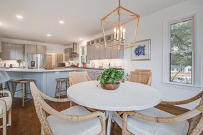 Uma cozinha moderna com uma mesa de jantar redonda cercada por cadeiras de vime, um lustre e vista para o exterior através de uma janela.