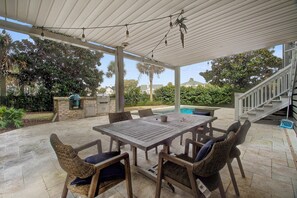 Um pátio externo com mesa e cadeiras, uma área coberta e uma piscina ao fundo.