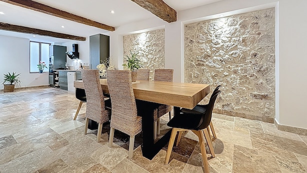 A dining area with a wooden table and woven chairs, a kitchen in the background, and a textured stone wall.