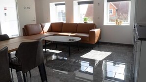 Ein moderner Lounge-Bereich mit einer Ledersofa, einem runden Tisch und einem Esstisch mit Stühlen. Zwei Fenster sorgen für natürliches Licht und bieten einen Ausblick nach draußen.
