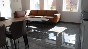 Ein modernes Zimmer mit einem braunen Ledersofa, einem runden Couchtisch und einem Esstisch mit Stühlen. Zwei Fenster sorgen für natürliches Licht und bieten einen Ausblick nach draußen.