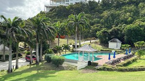 Un espace piscine agrémenté d’un kiosque, de chaises longues et d’un petit abri, le tout entouré de palmiers et bordé par une colline sur laquelle se trouve une maison au loin.
