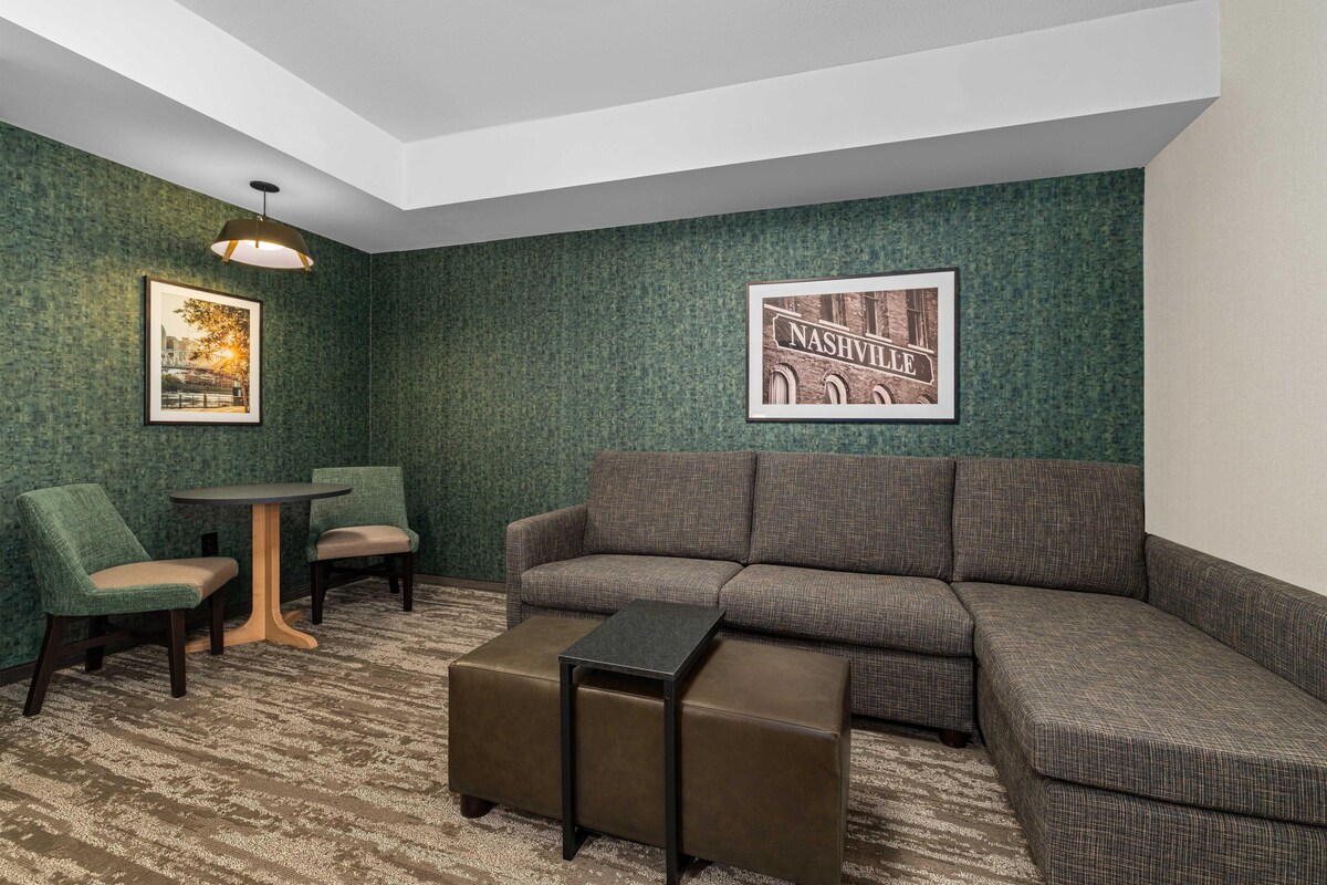 Quarto com sofá em formato de L, duas poltronas, uma mesinha redonda e uma mesa de centro. Há duas imagens emolduradas nas paredes.