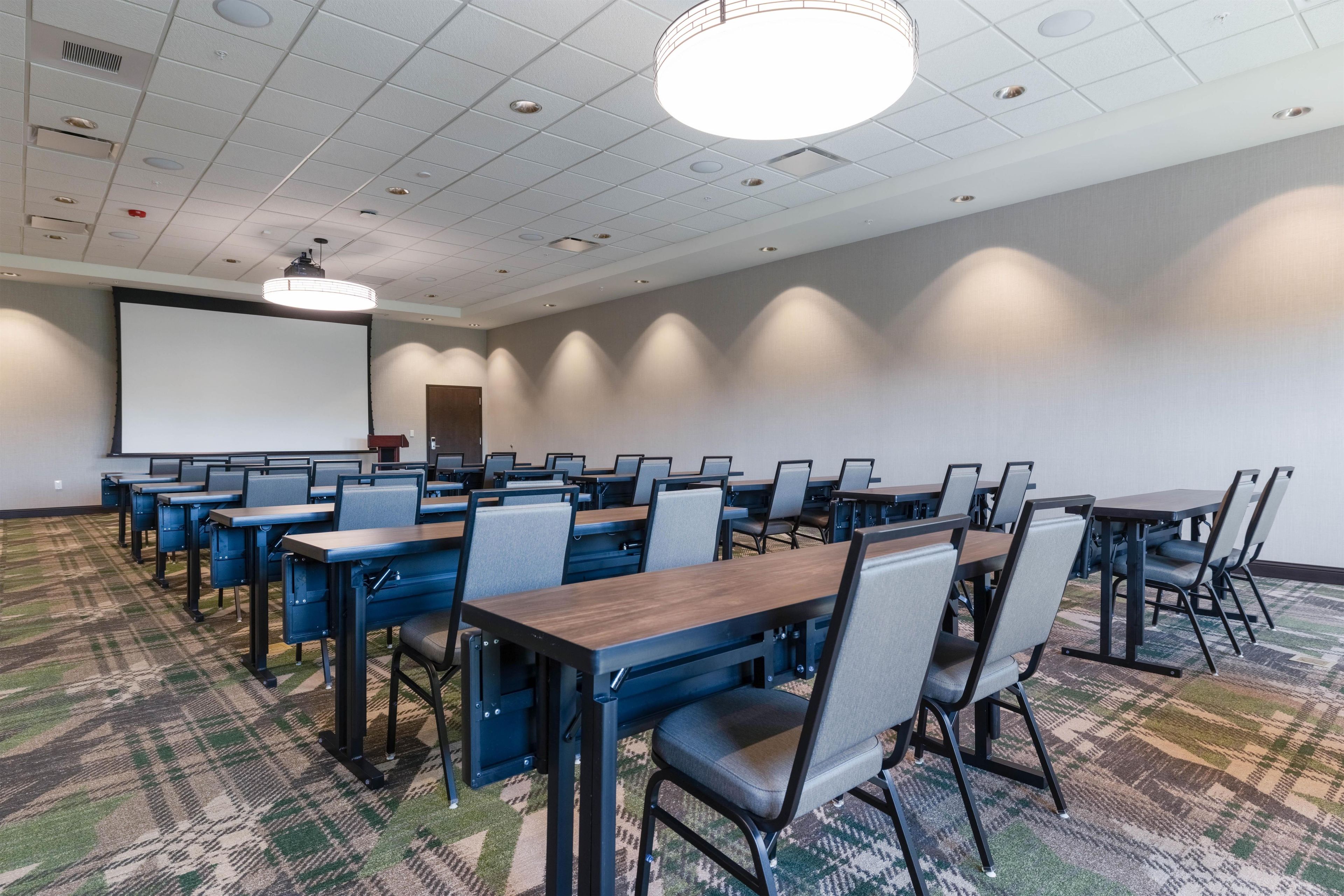 A conference room with multiple tables and chairs, a projector screen, and a carpeted floor.