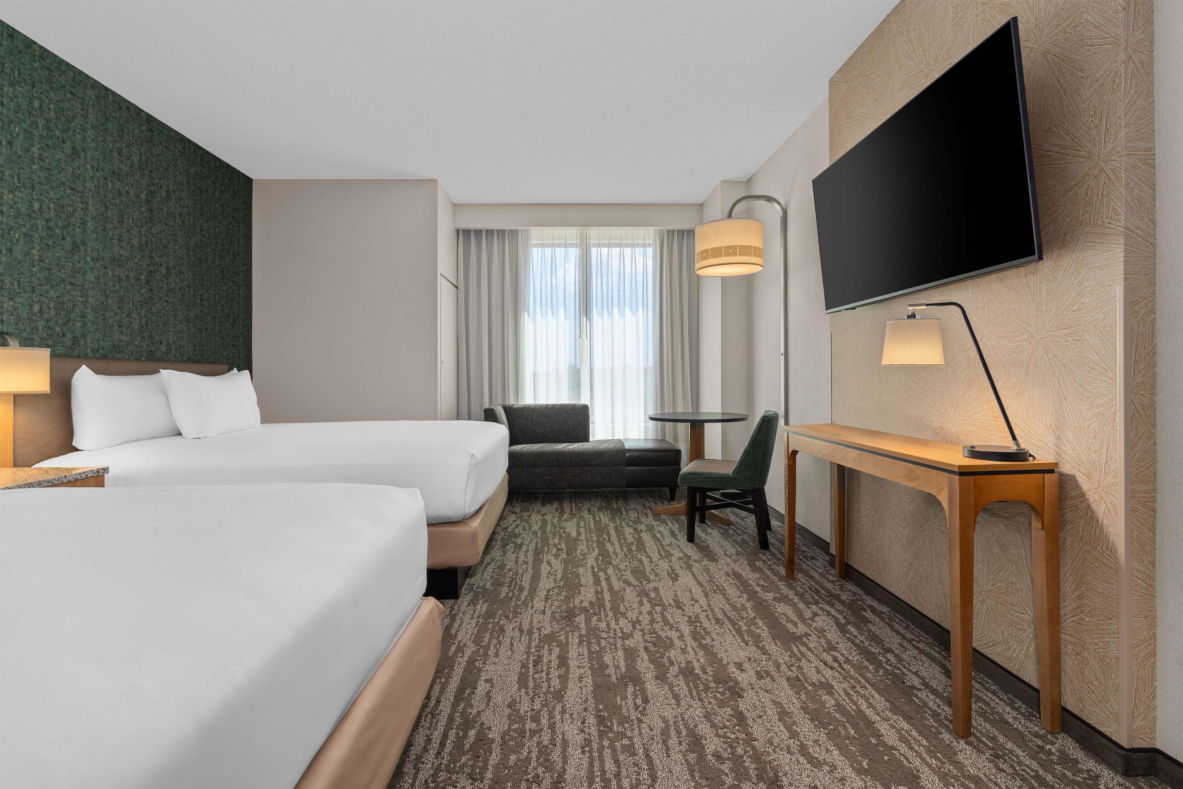 Ein Hotelzimmer mit zwei Betten, einem Schreibtisch, einem Sessel, einem Fernseher und einem Fenster mit Vorhängen.