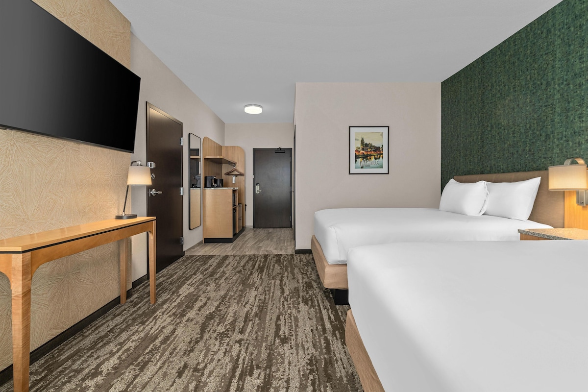 Quarto de hotel com cama, escrivaninha, espelho, guarda-roupa, porta e quadro na parede.