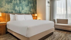 A hotel room with a large bed, a headboard with a forest-themed wallpaper, a side table with a lamp, a sofa, and a large window with curtains. - Drury Plaza Hotel Nashville Southeast Century Farms (Antioch)