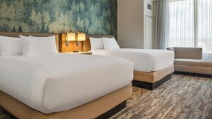 Habitación de hotel con dos camas, un ventanal con cortinas, una pared con estampado de bosque y un pequeño rincón de descanso.