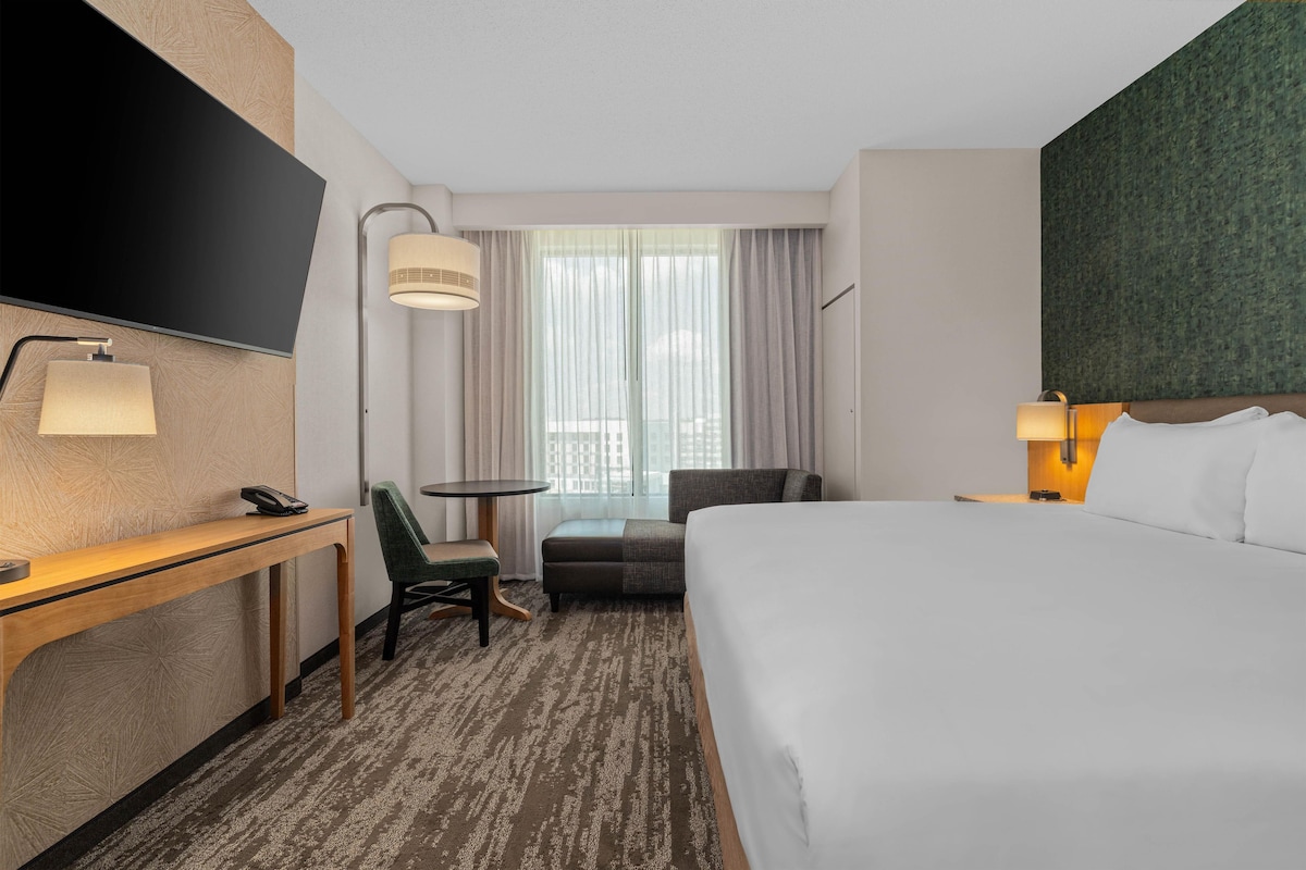 Quarto de hotel com cama, escrivaninha, cadeira, mesa pequena, luminária e janela com cortinas.