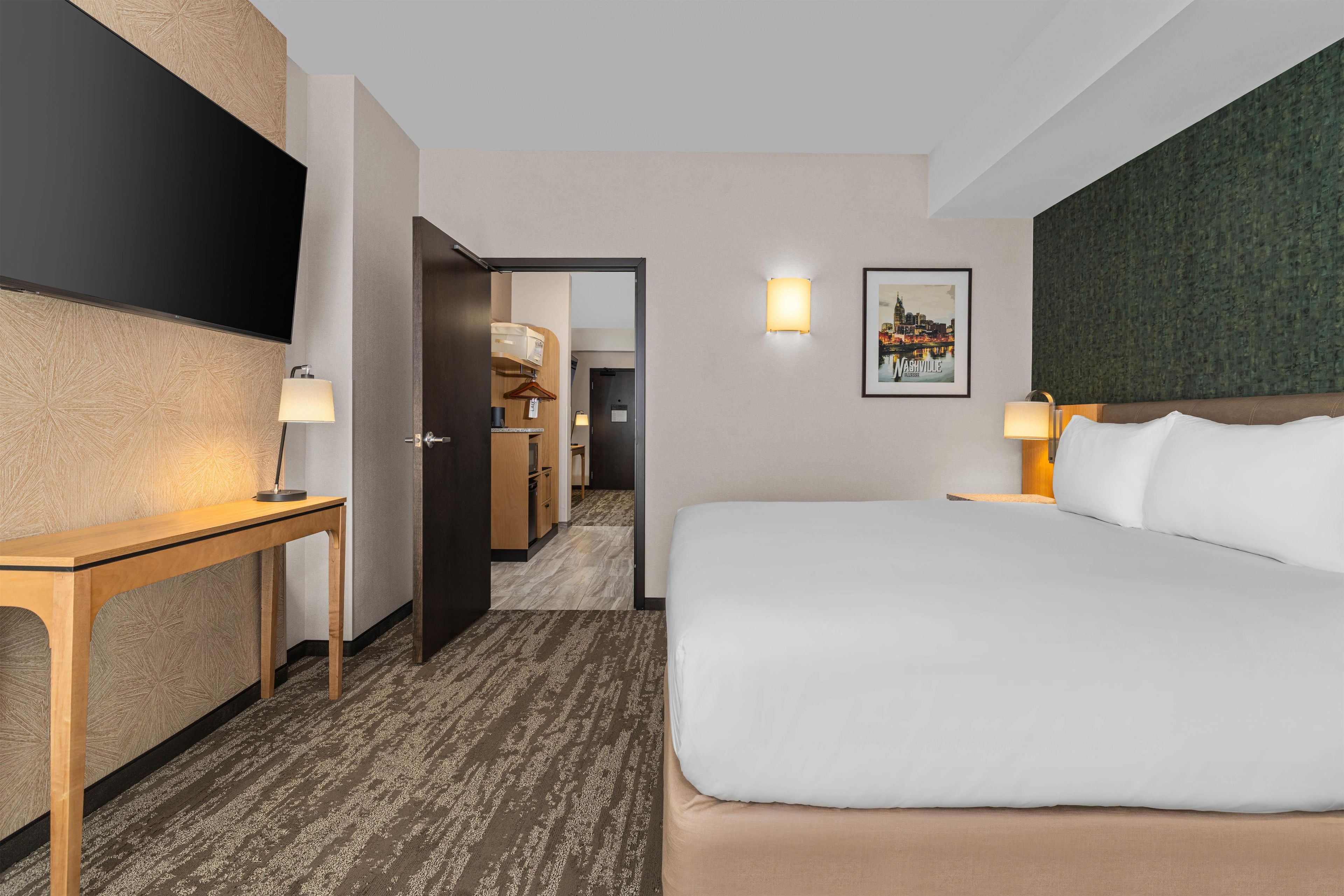 Ett hotellrum med en säng, ett skrivbord, en tv, en dörr som leder ut till en hall och en inramad tavla på väggen.